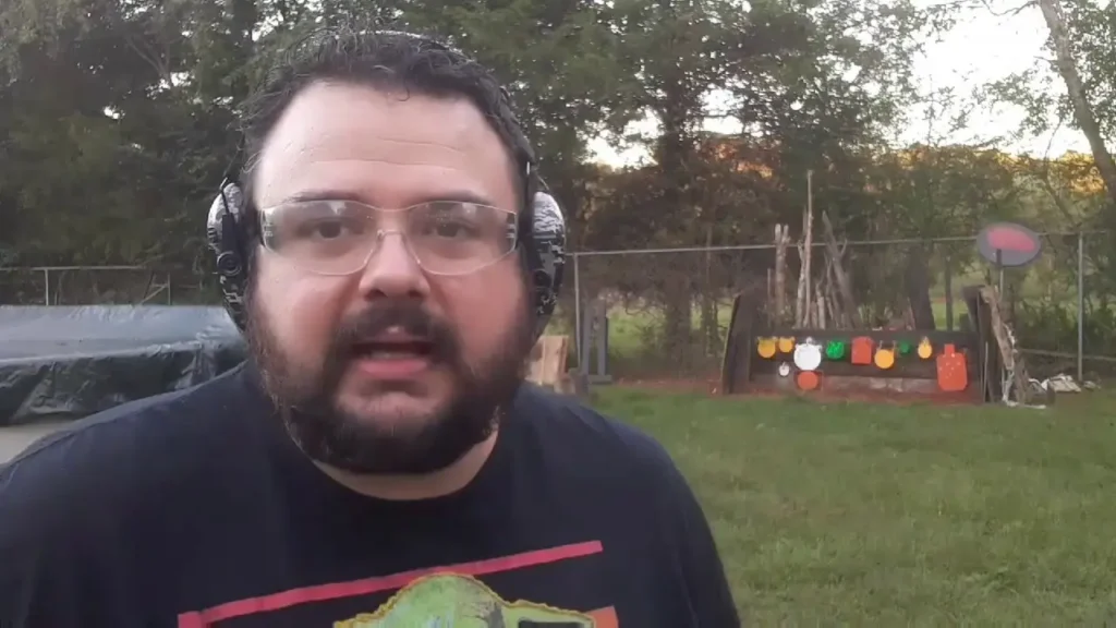 Man with a beard and safety glasses speaks to camera in a backyard shooting area with a colorful target board in the background.
