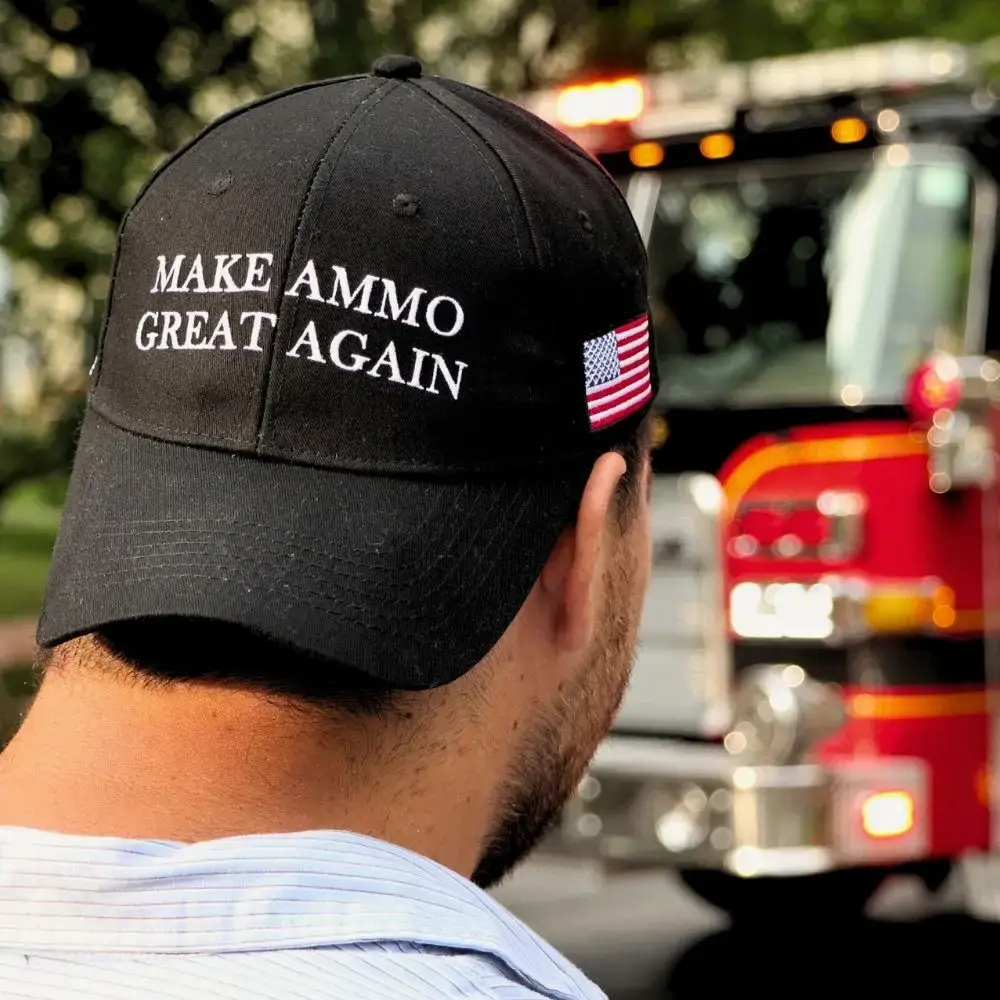Close-up of a man wearing a black cap that reads 'MAKE AMMO GREAT AGAIN' with a small American flag, blurred red fire truck in the background, outdoors.